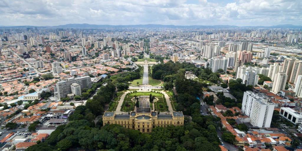 Vista aérea do Museu do Ipiranga em São Paulo, um edifício amarelo neoclássico cercado por jardins verdes e uma longa avenida com espelho dágua, em contraste com a vasta paisagem urbana de prédios altos e casas com telhados alaranjados sob um céu nublado