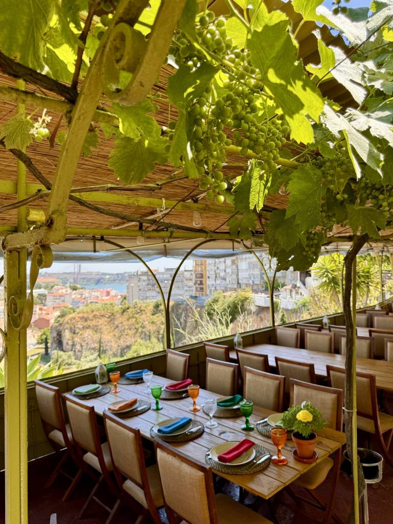 Terraço de restaurante com teto de palha e parreiras de uvas verdes, mesas de madeira postas com pratos, talheres, copos coloridos e guardanapos. Ao fundo, vista panorâmica de uma cidade com rio e ponte