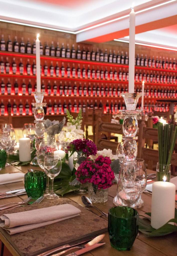 Mesa de jantar elegantemente posta com talheres, taças de vinho, guardanapos de tecido, velas acesas em castiçais de cristal e arranjos florais em tons de roxo e branco. Ao fundo, uma parede com prateleiras iluminadas em vermelho exibe centenas de garrafas de vinho