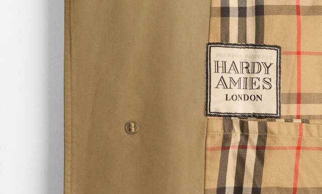 Detalhe de um casaco bege com forro xadrez em tons de bege, preto e vermelho. Uma etiqueta branca com borda preta, escrito HARDY AMIES LONDON, está costurada sobre o forro. Um botão bege com quatro furos aparece na parte bege do casaco