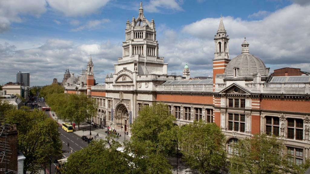 Vista aérea do Victoria and Albert Museum em Londres, com sua fachada de tijolos vermelhos e detalhes em pedra clara, incluindo uma torre central alta e uma cúpula. Uma rua arborizada com ônibus e carros passa em frente ao museu, sob um céu parcialmente nublado