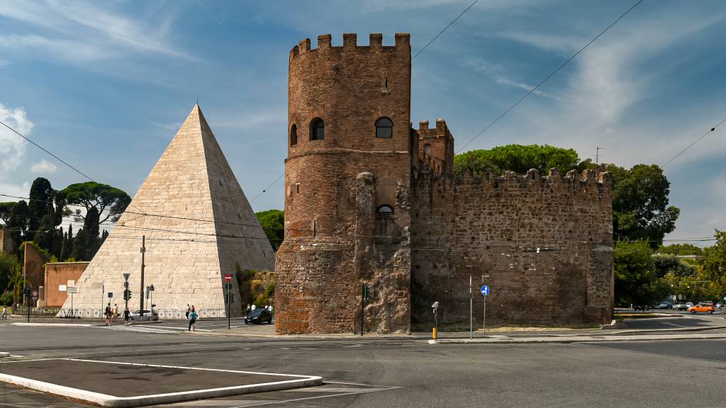 A Pirâmide de Céstio, monumento romano de pedra clara, e uma torre medieval de tijolos marrons com ameias, em uma rua movimentada sob céu azul com nuvens esparsas