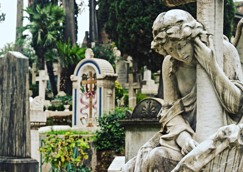 Estátua de anjo em mármore, com expressão de luto, apoiada em uma cruz, em um cemitério com túmulos e vegetação densa ao fundo