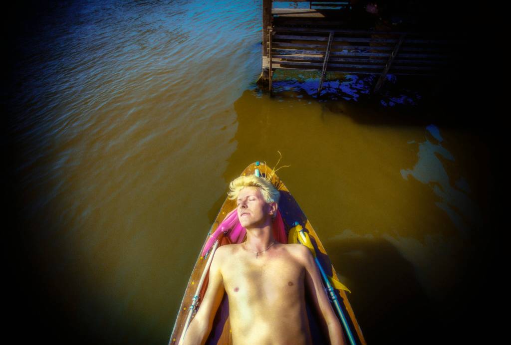 Homem de cabelo loiro platinado, sem camisa, deitado em um barco de madeira com detalhes coloridos, flutuando em águas escuras. Ele tem os olhos fechados e o rosto virado para cima, como se estivesse aproveitando o sol. Ao fundo, uma estrutura de madeira escura sobre a água. A imagem tem um contraste forte, com áreas claras e escuras acentuadas