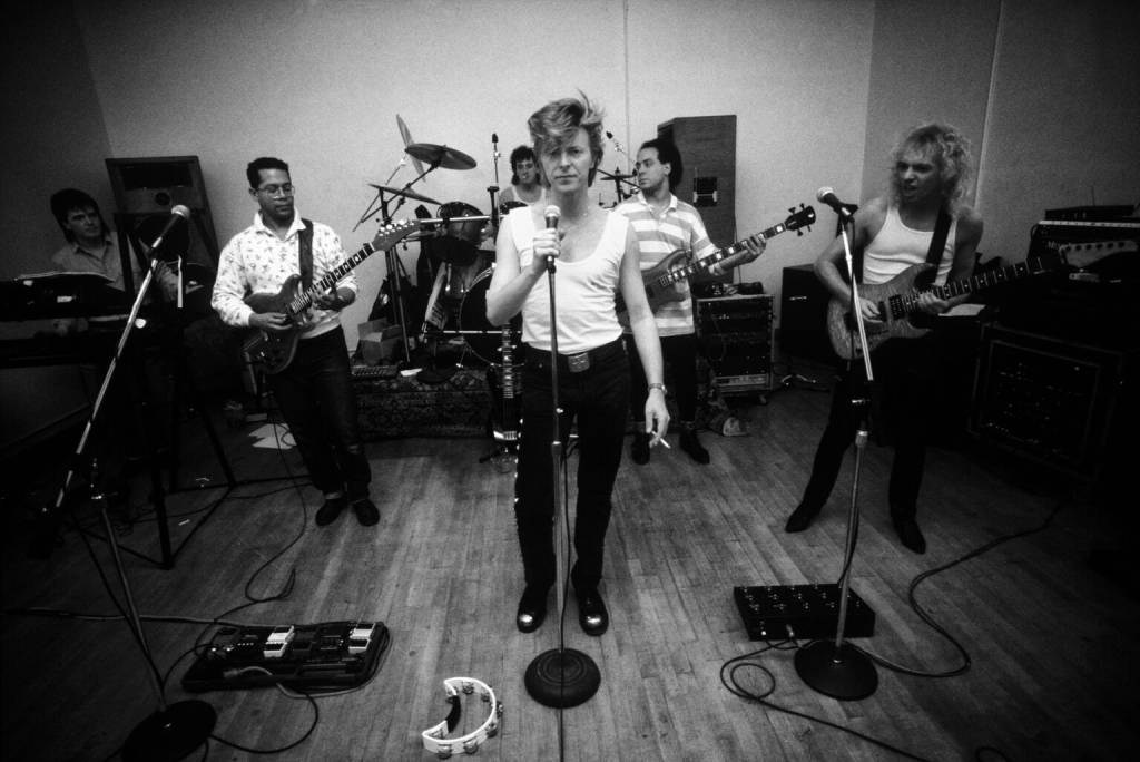 David Bowie com top branco e cabelo espetado, segurando um microfone, cercado por sua banda em um estúdio de ensaio. À esquerda, um guitarrista de camisa estampada e um tecladista. Atrás, um baterista. À direita, um baixista de camisa listrada e um guitarrista de regata. Pedais de efeito e um pandeiro estão no chão de madeira.