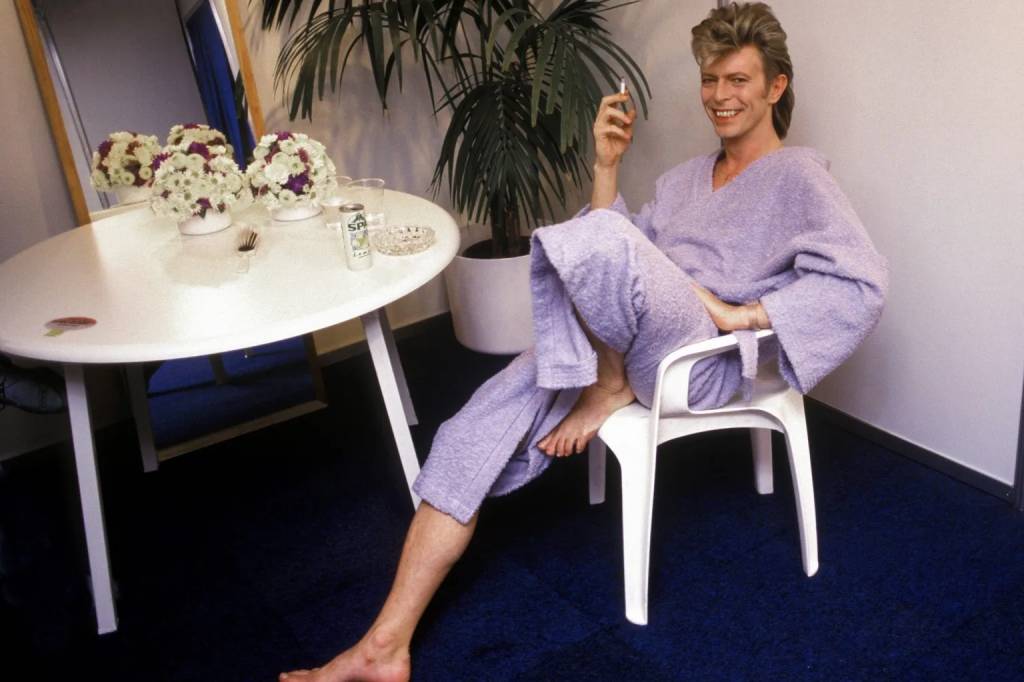 David Bowie sorridente, de roupão e calça lilás, descalço, sentado em cadeira branca, segurando um cigarro. À esquerda, uma mesa branca com arranjos florais, um cinzeiro e uma lata de refrigerante. Ao fundo, uma planta grande e um espelho. O chão é de carpete azul