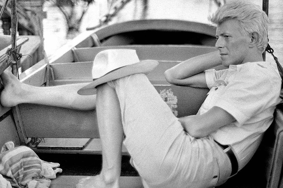 David Bowie, jovem, deitado descalço em um barco de madeira, com chapéu claro sobre a perna, camisa de manga curta e calça clara, olhando para a direita, com o braço esquerdo apoiado na borda do barco. A imagem é em preto e branco
