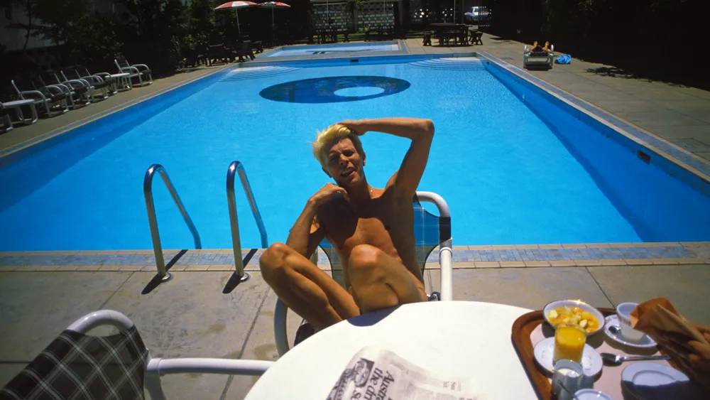 David Bowie sem camisa, com cabelo loiro espetado, sentado em uma cadeira branca ao lado de uma piscina azul. Ele olha para a câmera com um sorriso, a mão direita na cabeça e a esquerda no peito. Em primeiro plano, uma mesa branca com café da manhã e um jornal.
