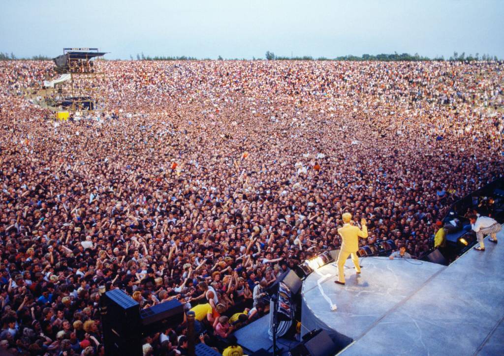 Um cantor de terno amarelo, de costas para a câmera, se apresenta em um palco curvo para uma multidão imensa em um festival ao ar livre. Outro músico agachado aparece à direita do palco. Ao fundo, uma torre de som e árvores distantes. O céu está claro
