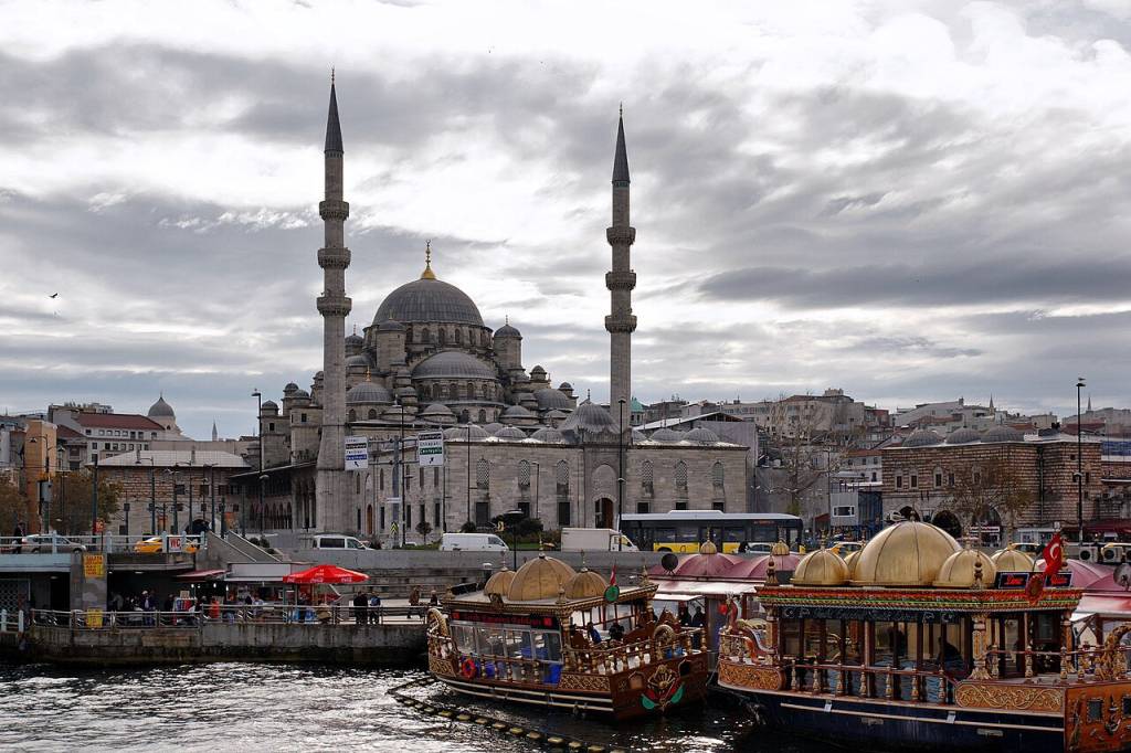 Mesquita Nova em Istambul, com dois minaretes altos e cúpulas cinzas, sob céu nublado. Em primeiro plano, barcos turísticos ornamentados com detalhes dourados flutuam em águas escuras, e um cais com pessoas e barracas vermelhas. Ao fundo, prédios da cidade e um ônibus amarelo na rua