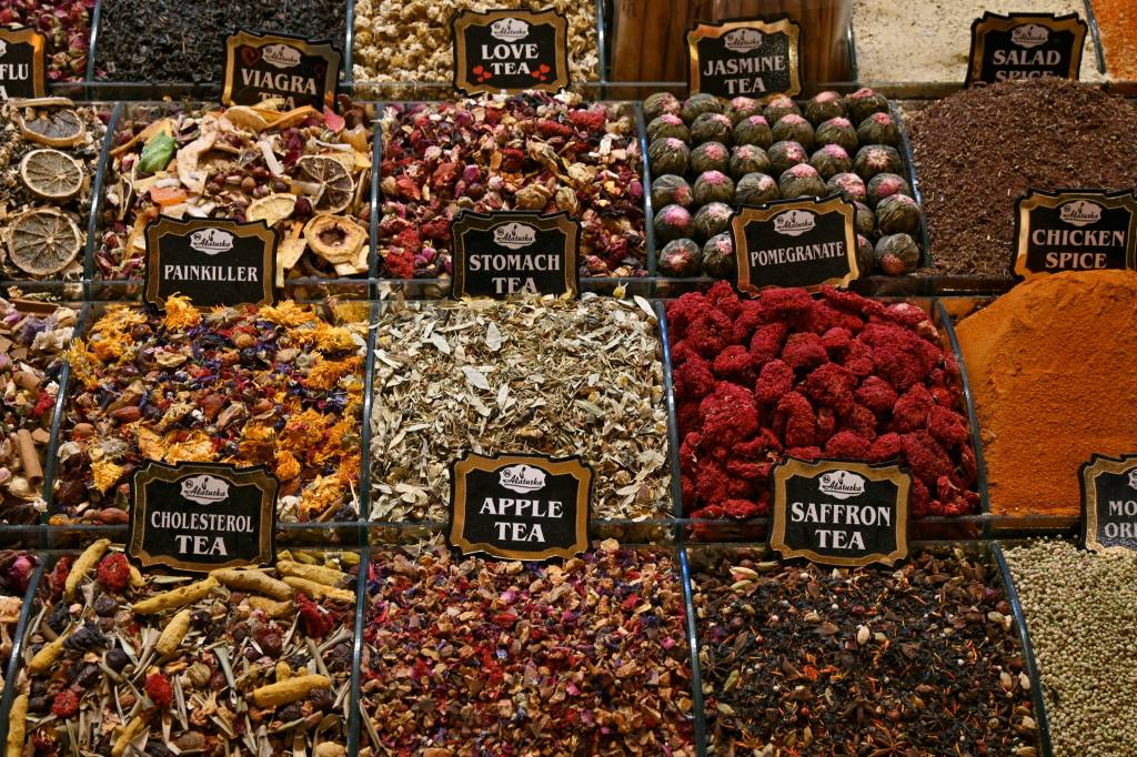 Exposição de chás e especiarias em um mercado, com diversas bandejas cheias de produtos coloridos e texturas variadas. Placas pretas com bordas douradas identificam os itens, como VIAGRA, PAINKILLER, CHOLESTEROL TEA, STOMACH TEA, APPLE TEA, SAFFRON TEA, LOVE TEA, JASMINE TEA, POMEGRANATE, SALAD SPICE e CHICKEN SPICE