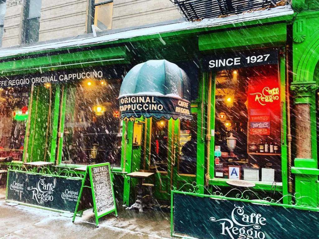 Fachada verde vibrante do Caffe Reggio em Nova York, com neve caindo. A placa SINCE 1927 e ORIGINAL CAPPUCCINO se destacam. Mesas externas e um quadro de menu de brunch estão cobertos por uma fina camada de neve, criando uma atmosfera aconchegante de inverno