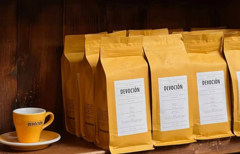 Prateleira de madeira escura com uma xícara de café amarela e pires à esquerda, e várias embalagens de café Devoción em pé, alinhadas à direita