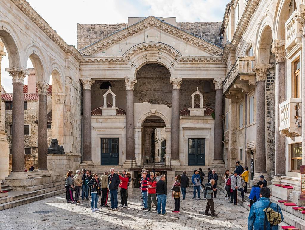 Turistas observam o peristilo do Palácio de Diocleciano, com colunas e arcos de pedra clara, sob um céu parcialmente nublado. Foto sob licença CC BY 2.0