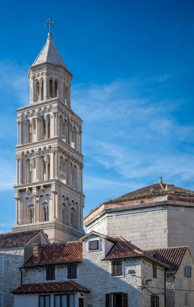 Campanário de pedra clara com telhado cônico e cruz no topo, em Split, Croácia. Abaixo, telhados de terracota e paredes de pedra de edifícios antigos, sob céu azul com nuvens esparsas