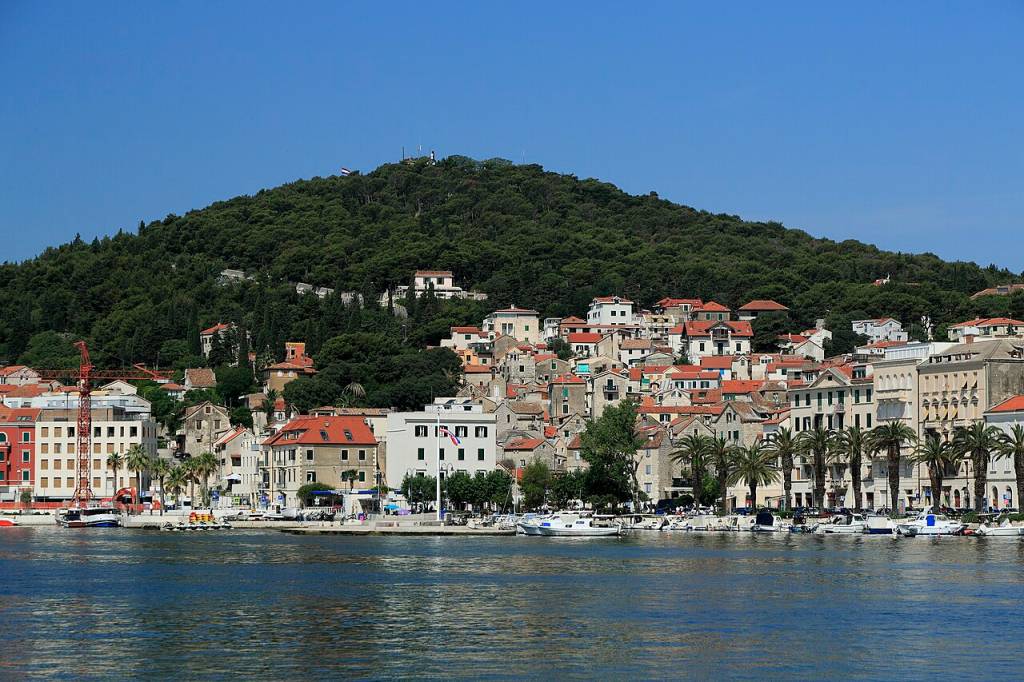 Vista panorâmica de uma cidade costeira com casas de telhados vermelhos e brancos, algumas com palmeiras, ao longo de um porto com barcos atracados, e uma montanha verdejante ao fundo sob céu azul claro