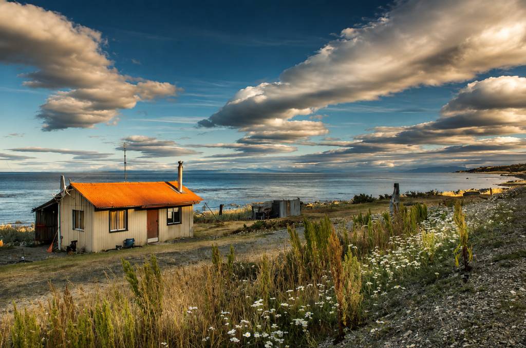 Uma pequena casa de madeira clara com telhado laranja e chaminé, à beira de um mar azul-escuro sob um céu com nuvens brancas e cinzas. A frente da casa tem uma porta marrom e duas janelas. Há vegetação rasteira e flores brancas em primeiro plano, e montanhas distantes no horizonte