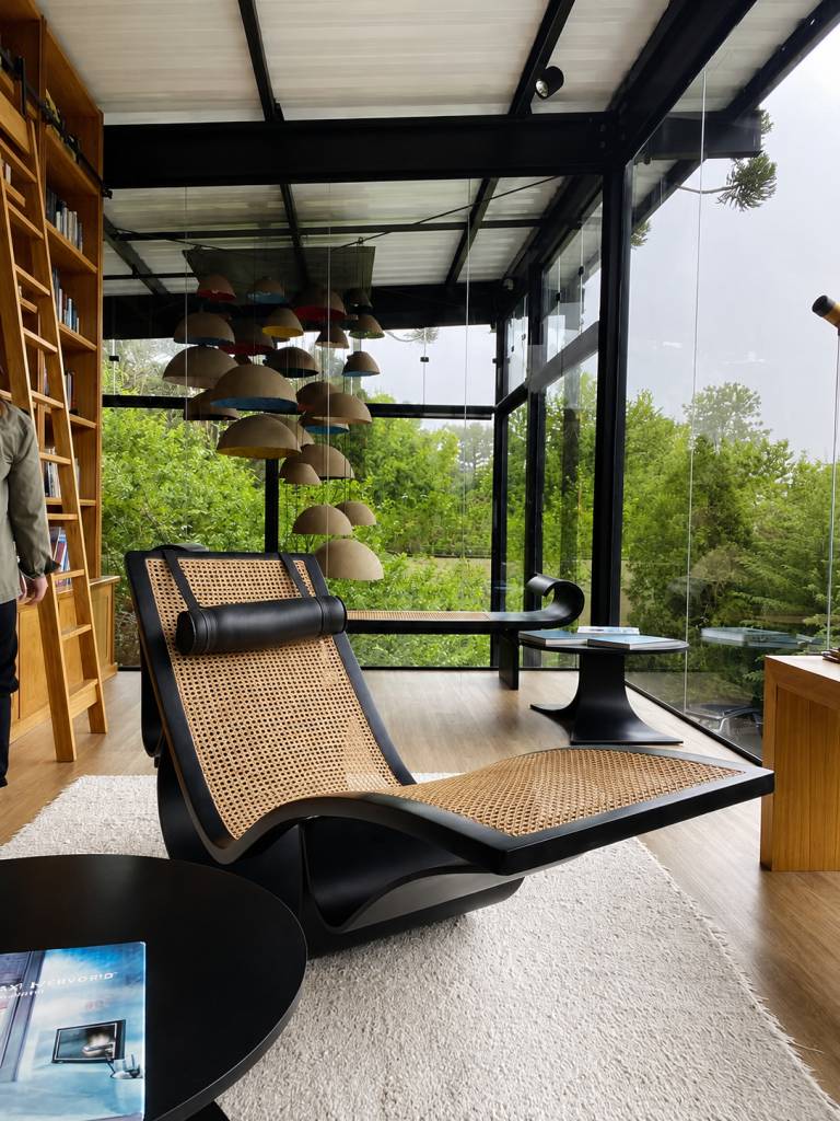 Ambiente moderno com poltrona reclinável preta e palha, estante de madeira com livros, e janelas do chão ao teto revelando uma paisagem verde. Um lustre com cúpulas de palha coloridas e uma mesa de centro preta com um livro completam a cena. Uma pessoa de jaqueta verde está à esquerda, e um telescópio é visível à direita