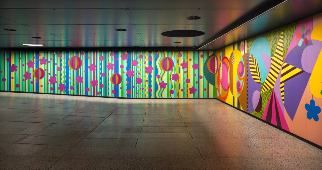 Corredor com paredes coloridas, à esquerda listras verticais verdes, azuis e amarelas com bolas listradas e flores magenta. À direita, formas geométricas abstratas em tons de roxo, amarelo, rosa e azul. O teto é escuro com luminárias embutidas e o chão é de lajotas cinzas