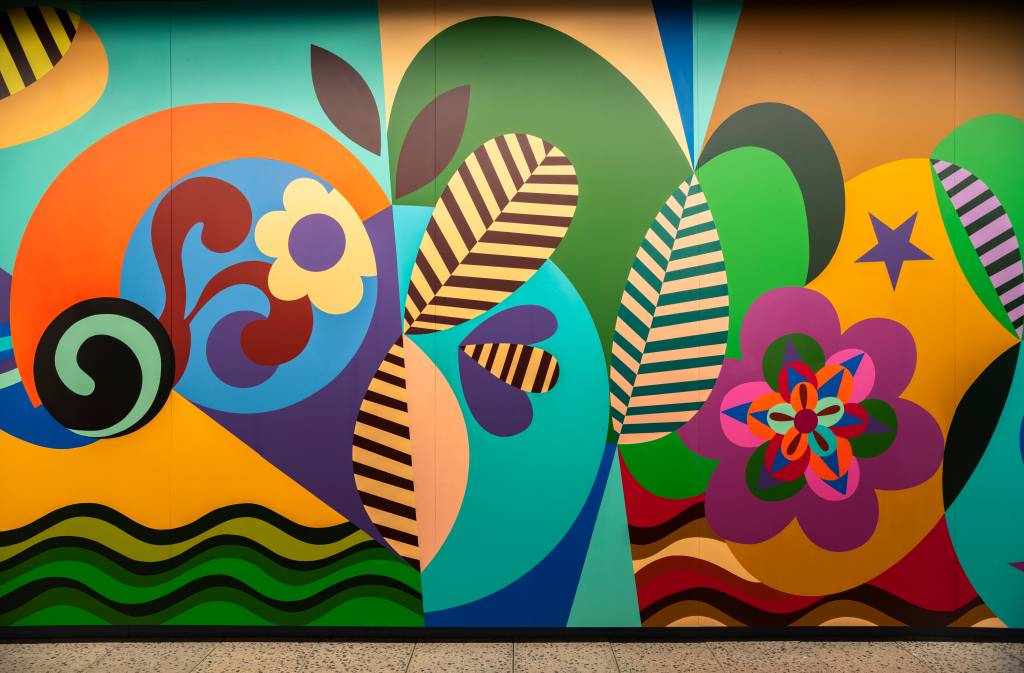 Mural colorido com formas abstratas e orgânicas em tons vibrantes de azul, verde, laranja, roxo e amarelo. Destacam-se flores estilizadas, folhas listradas e ondas, criando um padrão dinâmico e alegre