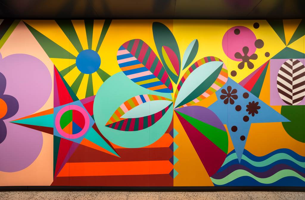 Mural vibrante com formas geométricas e orgânicas em cores fortes como amarelo, azul, rosa, verde e laranja. Elementos incluem um sol, folhas listradas, flores e ondas, criando uma composição abstrata e alegre