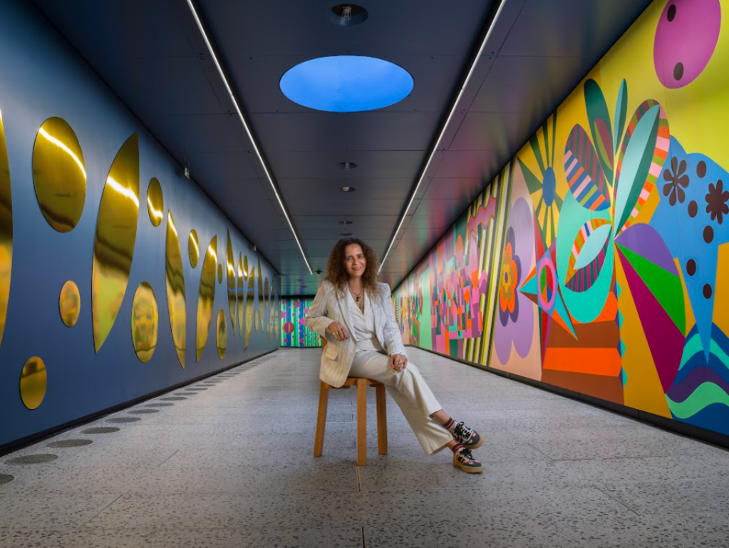 Mulher de cabelos cacheados, vestindo blazer e calça brancos, sentada em um banco de madeira em um corredor com paredes artísticas. À esquerda, painéis dourados refletem a luz; à direita, um mural vibrante com formas geométricas e florais coloridas. Um círculo azul no teto ilumina o ambiente