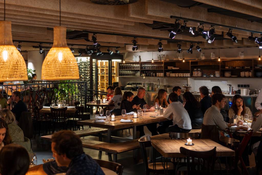 Pessoas sentadas em mesas de madeira rústica em um restaurante com iluminação aconchegante, lustres de palha e prateleiras com garrafas de vinho ao fundo