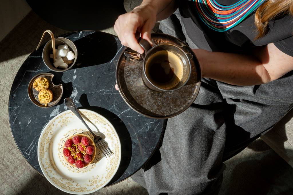 Visão superior de uma pessoa segurando uma xícara de café sobre um pires metálico, sentada à mesa de mármore preto com doces. Na mesa, há um prato com uma tortinha de framboesa e pistache, um garfo, um recipiente com cubos de açúcar e pinça, e outro com duas mini tortinhas. A pessoa veste uma blusa preta e calça cinza, com um colar colorido. A luz solar incide sobre a cena, criando sombras.