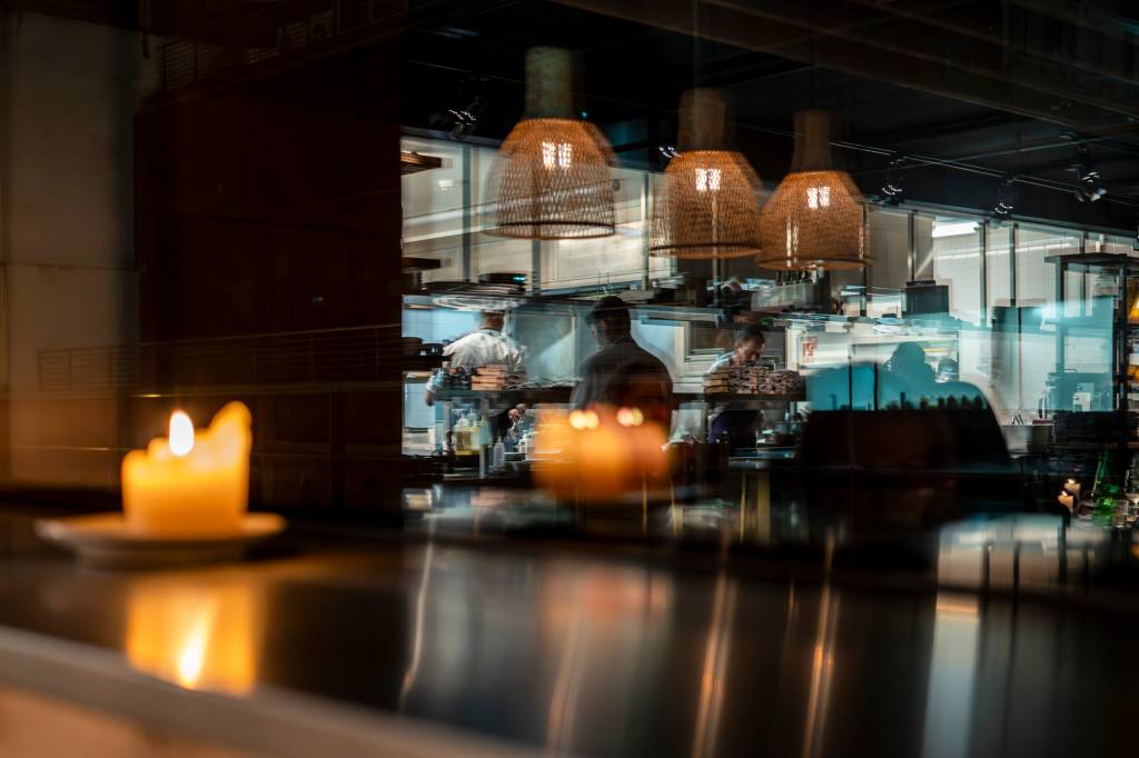 Reflexo de uma cozinha de restaurante com três chefs trabalhando, vista através de um balcão escuro com duas velas acesas em primeiro plano, criando um ambiente intimista
