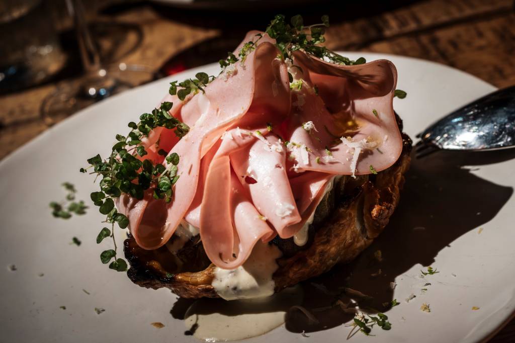 Prato gourmet com fatias de mortadela dobradas, queijo ralado, azeite e brotos verdes sobre uma base crocante, com molho cremoso escorrendo, em um ambiente com luz baixa
