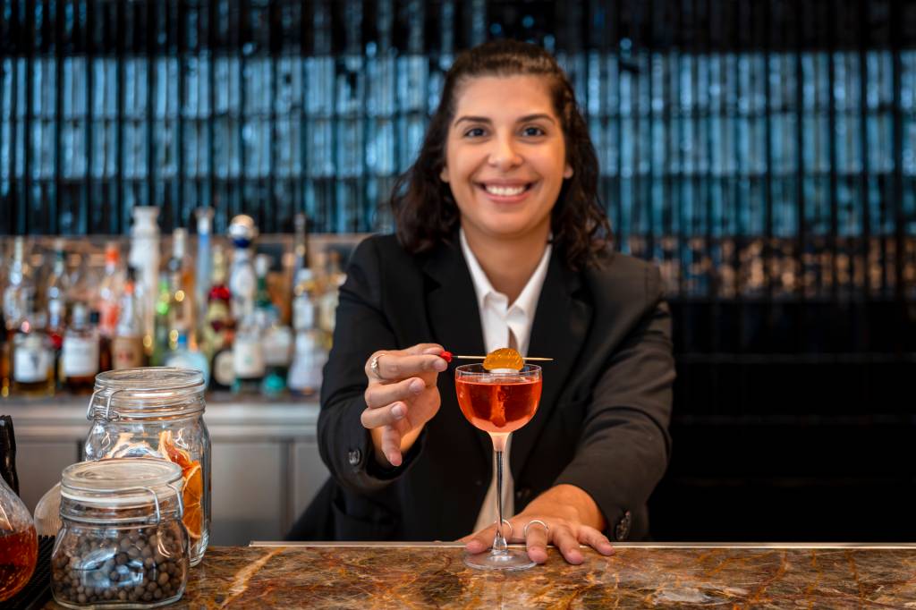 Uma mulher sorridente, de cabelos escuros e blazer preto, segura um palito com uma framboesa sobre um coquetel avermelhado em um balcão de bar. Garrafas de bebidas e potes de temperos estão ao fundo