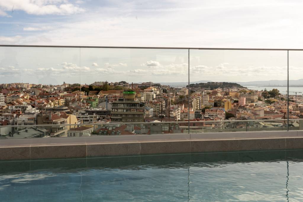 Vista panorâmica de uma cidade com telhados alaranjados e edifícios coloridos, com um rio ou mar ao fundo, sob um céu parcialmente nublado. Em primeiro plano, uma piscina de borda infinita e um parapeito de vidro transparente.