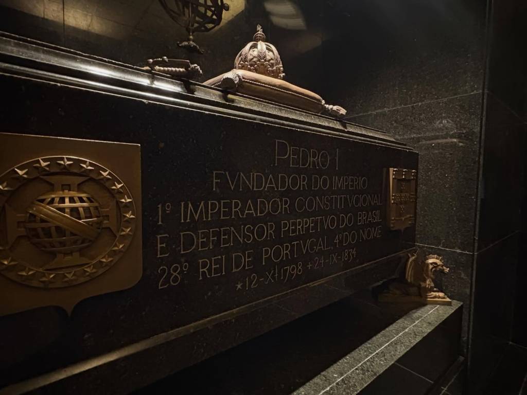 Túmulo escuro de mármore com a inscrição PEDRO I FVNDADOR DO IMPERIO 1º IMPERADOR CONSTITVCIONAL E DEFENSOR PERPETVO DO BRASIL 28º REI DE PORTVGAL, 4º DO NOME *12-X-1798 +24-IX-1834. À esquerda, um brasão dourado com esfera armilar e estrelas. No canto inferior direito, uma pequena escultura de leão dourado. O ambiente é escuro, com iluminação focada no túmulo