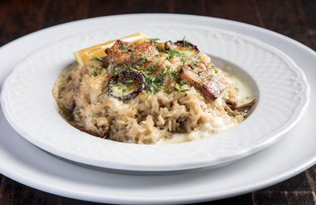 Risoto cremoso com pedaços de carne grelhada, cogumelos e queijo derretido, decorado com salsa picada e uma fatia de limão, servido em prato branco sobre fundo escuro