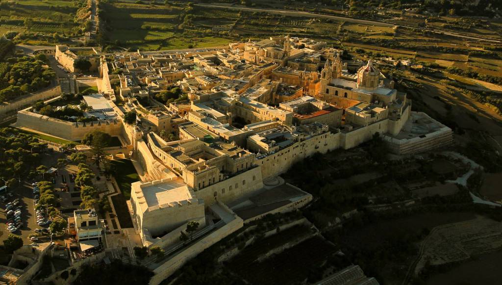 Vista aérea de Mdina, Malta, uma cidade fortificada com edifícios de pedra clara e telhados planos, cercada por muralhas e campos verdes. A luz do sol poente ilumina as construções, criando sombras longas e um brilho dourado. Uma grande cúpula e torres de igreja se destacam no centro da cidade, com um estacionamento visível à esquerda.