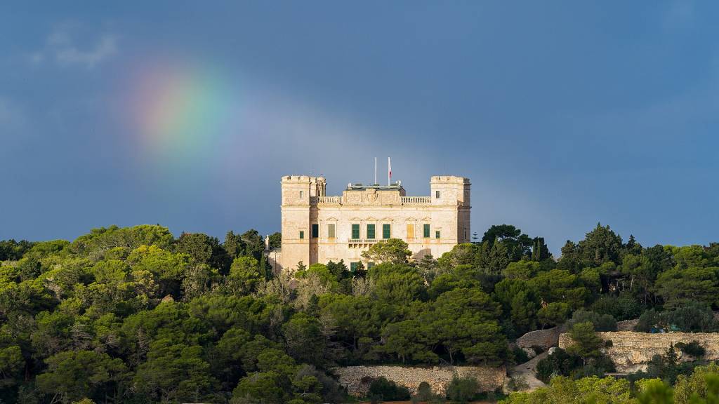 Castelo histórico de pedra clara com torres e janelas verdes, cercado por densa floresta verde sob um céu azul-escuro com um arco-íris visível à esquerda