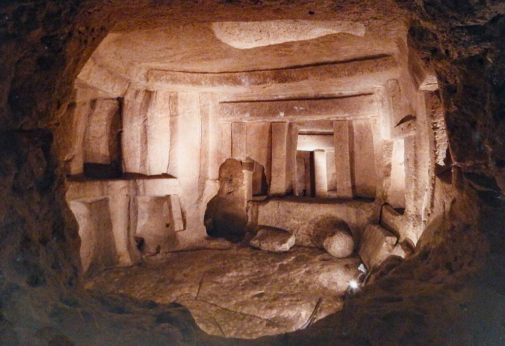 Interior de uma tumba etrusca escavada na rocha, com paredes e teto irregulares em tons de marrom e bege. Há nichos e saliências esculpidas, e no fundo, uma abertura retangular escura, sugerindo outra câmara. O chão é de terra batida com algumas pedras soltas, e a iluminação é difusa, criando sombras profundas