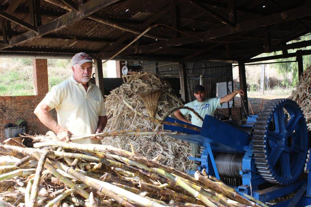 Dois homens em um galpão rústico com telhado de zinco. Um homem de boné e camisa polo creme segura cana-de-açúcar, olhando para frente. Outro homem de boné e camiseta clara opera uma máquina azul com engrenagens, ao lado de um monte de bagaço de cana. Há um monte de cana-de-açúcar cortada em primeiro plano