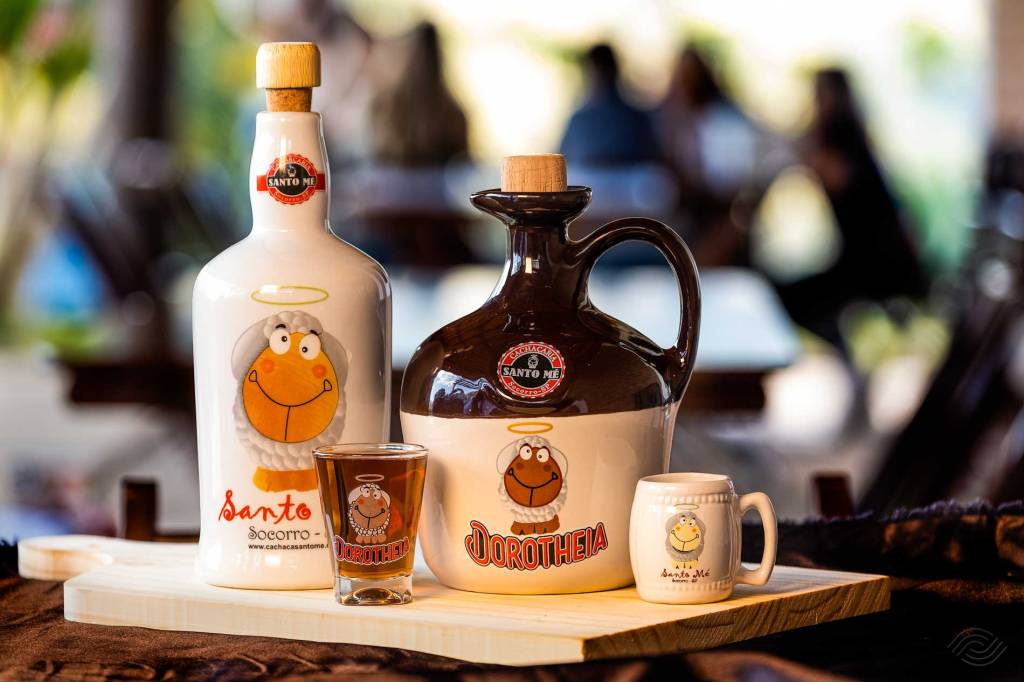 Garrafa branca de cachaça Santo Mé Santo Socorro com ovelha sorridente, copo de shot Dorotheia e caneca branca com ovelha, sobre uma tábua de madeira