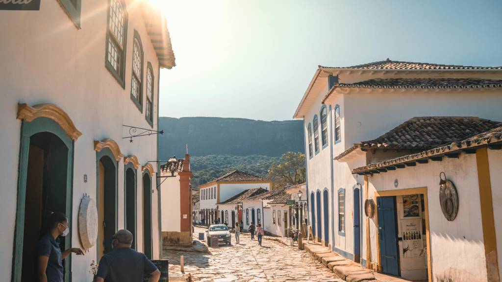 Rua de paralelepípedos em cidade histórica, com casarões coloniais brancos e detalhes coloridos. O sol brilha forte no céu azul, iluminando as fachadas e as montanhas ao fundo. Pessoas caminham pela rua, e um carro está estacionado.