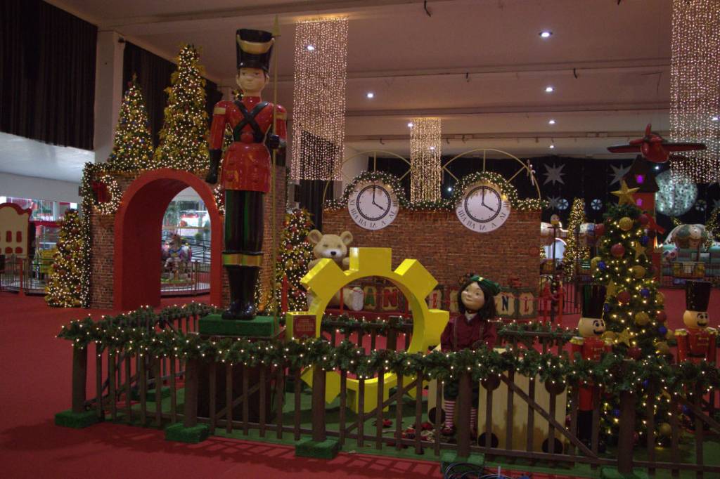 Decoração natalina com soldadinho de chumbo gigante, árvores de Natal iluminadas, relógios de parede e um urso de pelúcia em um cenário festivo com luzes e enfeites