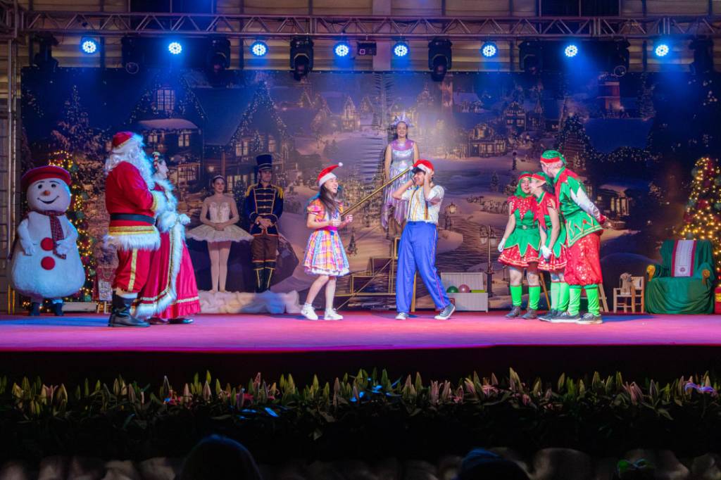 Elenco de Natal em um palco com fundo de vilarejo nevado. Da esquerda, um boneco de neve, Papai Noel e Mamãe Noel, uma bailarina, um soldadinho de chumbo, uma menina de gorro e vestido xadrez, um menino de gorro e suspensórios, uma fada e dois duendes. O palco é iluminado por luzes azuis e há plantas na frente