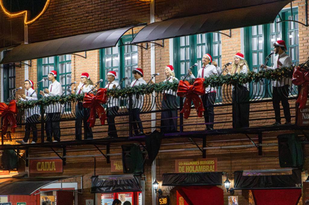 Um coral de 10 pessoas, vestindo camisas brancas, gravatas vermelhas e gorros de Papai Noel, canta em uma sacada decorada com festões e laços vermelhos. Ao fundo, uma parede de tijolos e janelas verdes. Abaixo, letreiros comerciais e a frase OS MELHORES BURGERS DE HOLAMBRA!. A cena noturna é iluminada por luzes festivas