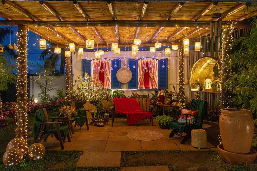 Pátio externo decorado com luzes de Natal, incluindo um pergolado de bambu iluminado, janelas azuis com cortinas vermelhas e um banco de madeira com manta vermelha. Há cadeiras, plantas e vasos de cerâmica, criando um ambiente festivo e acolhedor à noite