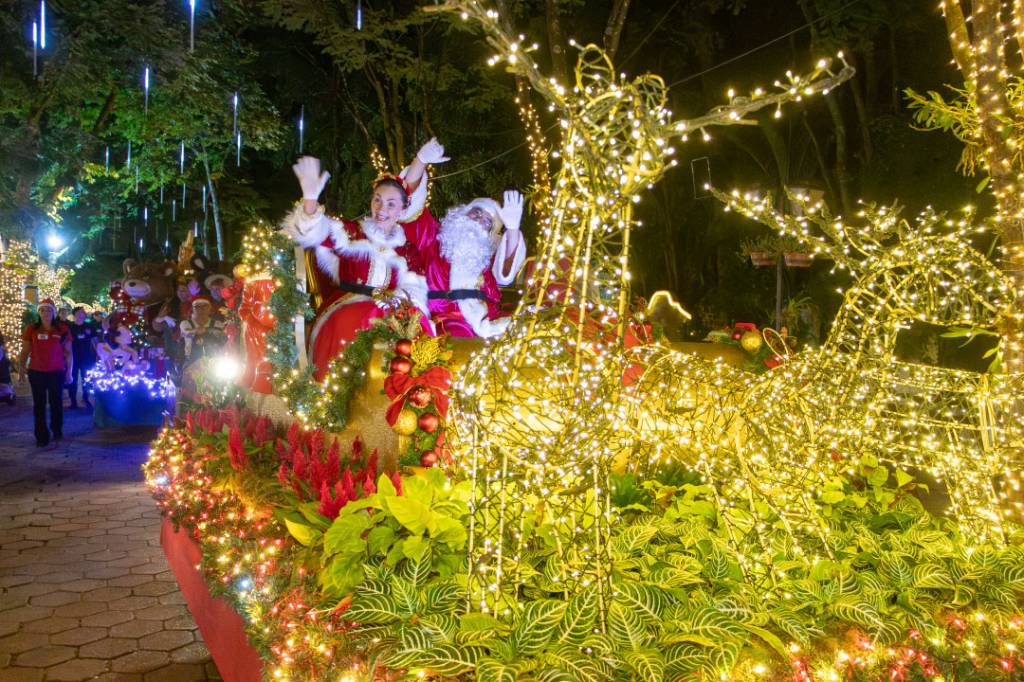 Papai Noel e Mamãe Noel sorridentes acenam de um trenó iluminado, puxado por renas de luz, em um desfile noturno de Natal. O cenário é um parque com árvores decoradas e pessoas ao fundo, criando uma atmosfera festiva e mágica