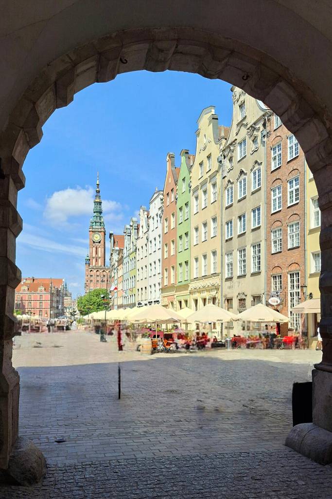 Vista através de um arco de pedra para uma rua de paralelepípedos com edifícios coloridos de telhados pontiagudos e um relógio alto ao fundo. Mesas de café com guarda-sóis bege ocupam a calçada, sob céu azul claro