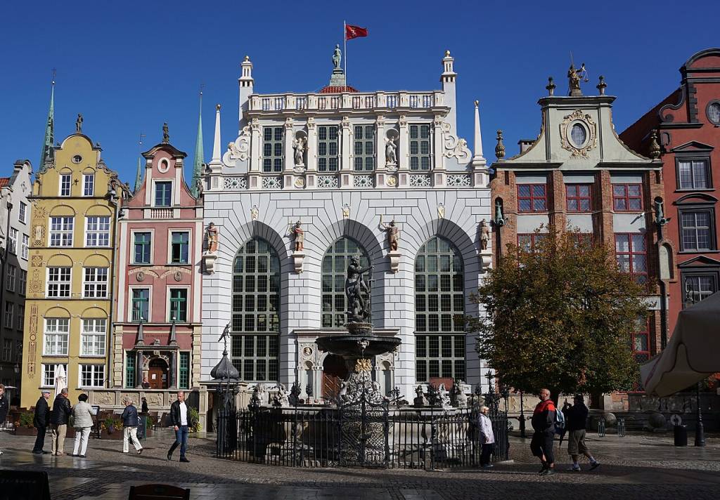 Edifícios históricos coloridos em Gdansk, Polônia, com uma fonte ornamentada em primeiro plano. O prédio central, branco, tem três grandes arcos e estátuas. À esquerda, edifícios amarelos e rosados; à direita, um prédio marrom-avermelhado. Pessoas caminham pela praça ensolarada. Uma árvore com folhagem outonal está à direita da fonte.