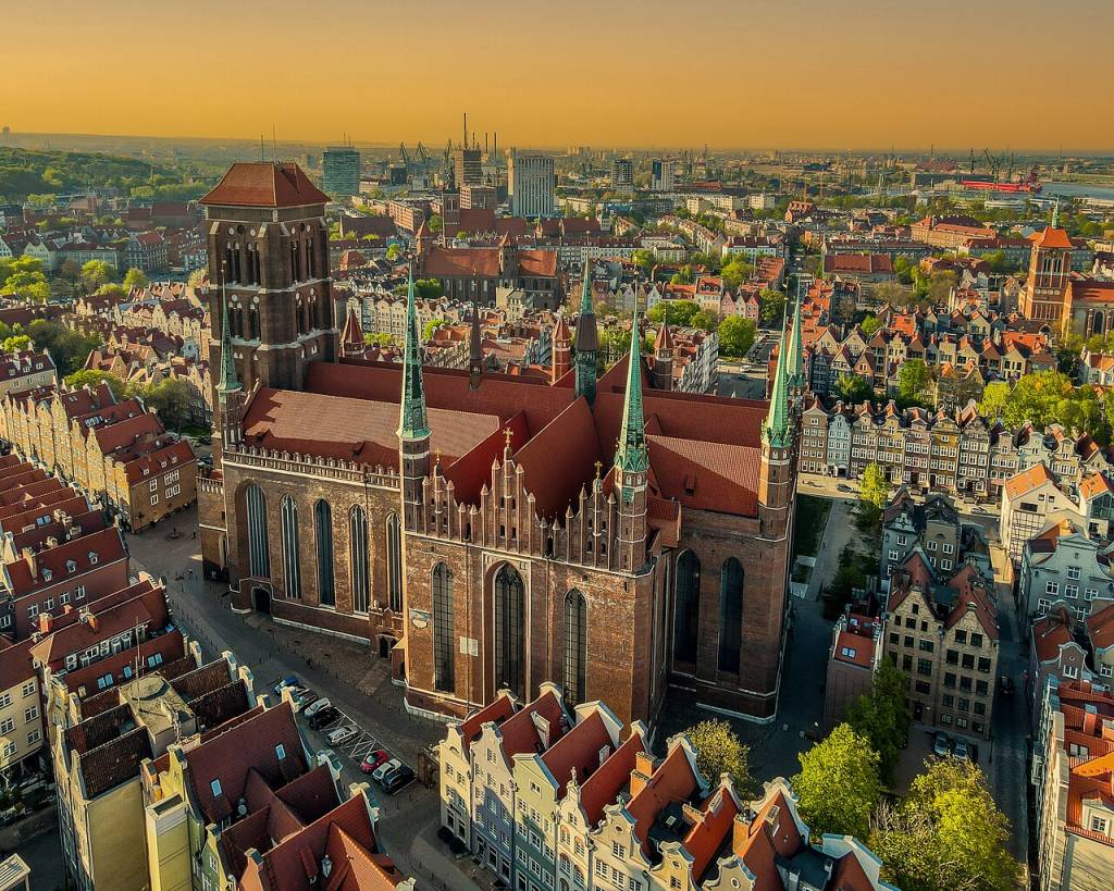 Vista aérea da Basílica de Santa Maria em Gdansk, Polônia, com telhados de terracota e torres verdes pontiagudas, cercada por edifícios históricos e uma paisagem urbana extensa sob um céu alaranjado ao pôr do sol