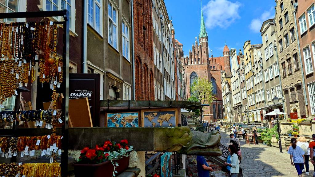 Rua de paralelepípedos em Gdansk, Polônia, com barracas de âmbar à esquerda, prédios históricos coloridos e uma igreja gótica ao fundo, sob céu azul com nuvens brancas. Pessoas caminham pela rua movimentada