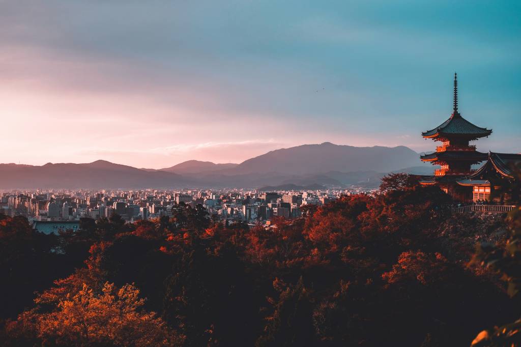 Vista panorâmica de uma cidade japonesa ao entardecer, com uma pagoda tradicional de telhados curvos e pontiagudos à direita, em meio a árvores com folhagem avermelhada. Ao fundo, montanhas e o céu em tons de azul e rosa.